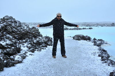 Full length of man with arms outstretched by rock formation at sea shore during winter