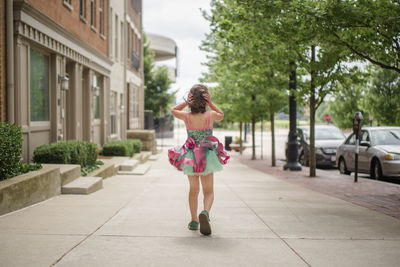 A small girl walks down sidewalk in tutu holding hair against wind