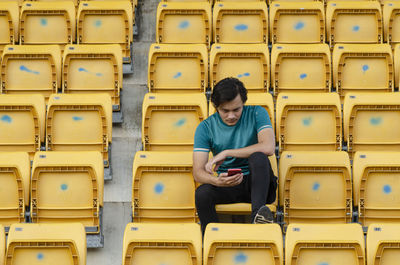 Smiling young man using phone sitting on chair in stadium