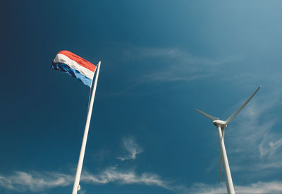 Low angle view of wind turbine and dutch flag against blue sky