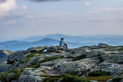 Rear view of man sitting on rock against mountains and sky