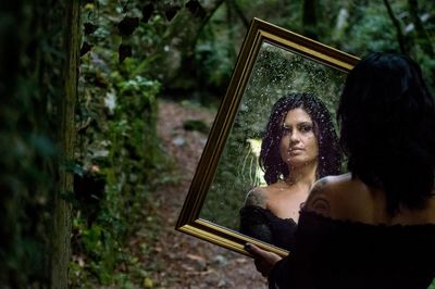 Rear view of beautiful woman holding wet mirror with reflection in forest