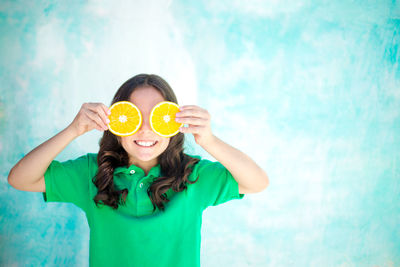 Happy girl holding orange slices in front of eyes while standing by turquoise wall