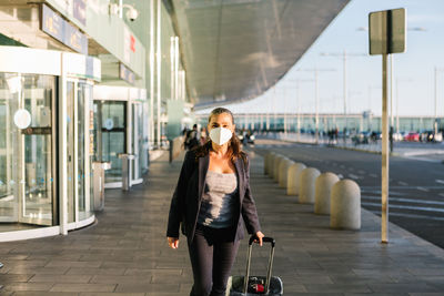 Cheerful female tourist in mask with suitcase and backpack standing in airport and smiling at camera while enjoying summer weekend