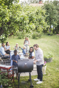 High angle view of man preparing barbecue grill with boy while family and friends sitting at table in backyard
