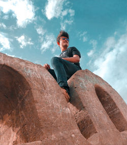 Low angle view of young man sitting on rock against sky