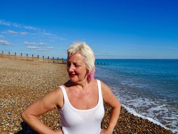 Smiling mature woman with hands on hip standing at beach against blue sky during sunny day