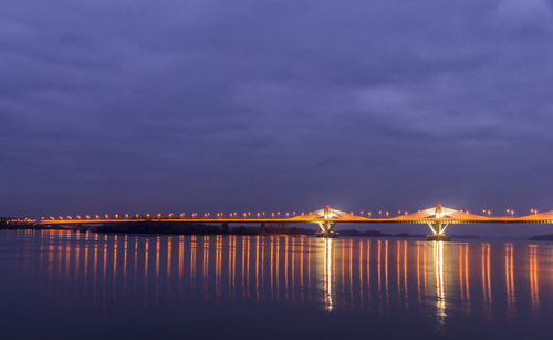 Illuminated bridge over sea against cloudy sky at dusk