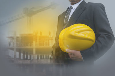 Digital composite image of architect holding hardhat while standing at construction site