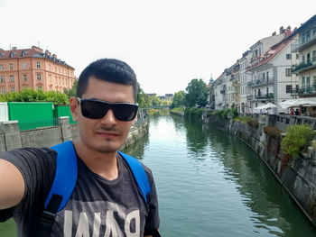Portrait of man wearing sunglasses standing by canal against clear sky
