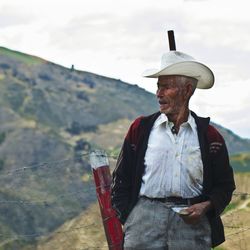 Senior man wearing cowboy hat standing by fence against sky