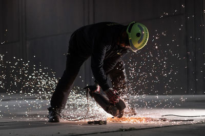 Full body of unrecognizable male welder in protective helmet and goggles cutting iron details with chop saw during work at industrial factory at night