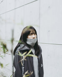 Close-up portrait of young woman covering mouth with turtleneck against wall