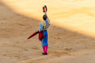 Back view of unrecognizable bullfighter in fancy costume taking off hat after corrida performance while standing on sandy arena