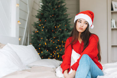 Young sad asian woman in cozy red sweater and santa hat on bed in room with christmas tree at home