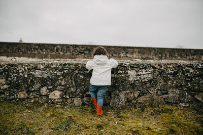 Rear view of girl standing by stone wall against sky