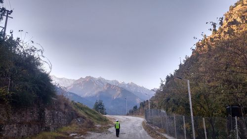 Rear view of man walking on road leading towards mountains against clear sky