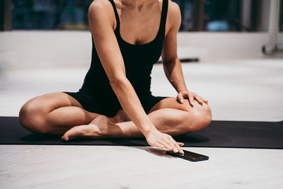 Low section of young woman sitting on mat while using mobile phone at gym