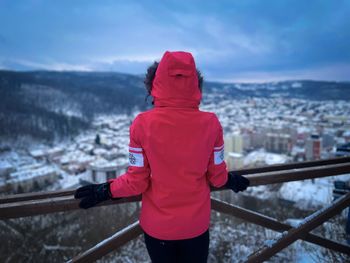 Rear view of woman with red hooded coat leaning against a wooden railing and looking at the city