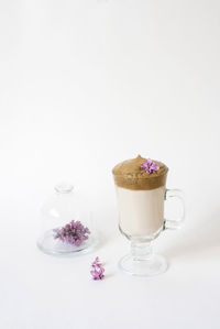 Dalgona coffee in a transparent mug with a lilac flower on a white background and a sprig 