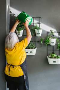 Back view of anonymous elderly female in casual clothes standing near shelves with potted plants and taking care of potted plants