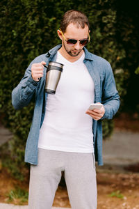 Man holding coffee cup while using mobile phone standing against trees