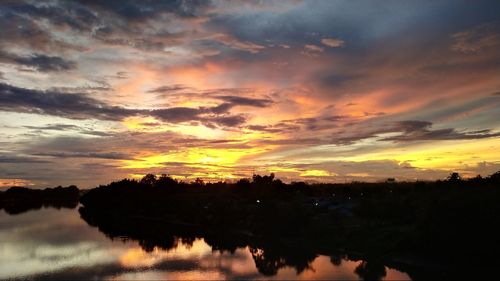 Silhouette trees by lake against dramatic sky during sunset