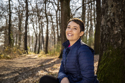 Young smiling woman sits at the foot of an oak tree in the woods.