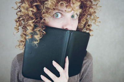 Portrait of shocked young woman covering mouth with book against gray background
