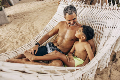 Shirtless father and son lying down in hammock at beach on vacation