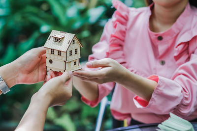 Midsection of girl holding model house with friend while sitting against plants