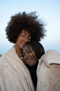 Happy young multiracial female best friends wrapped in warm blanket cuddling with eyes closed while chilling and enjoying time together in summer evening during holidays on seashore