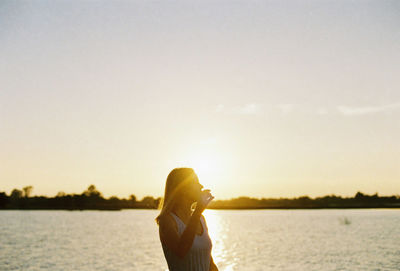 Side view of woman standing by lake against sky during sunset