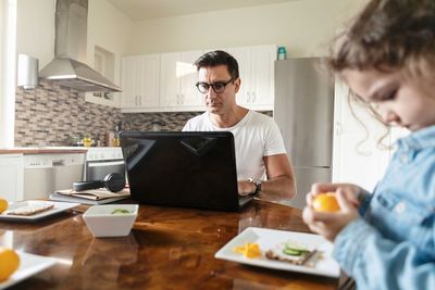 Daughter peeling orange fruit while father working over laptop at home