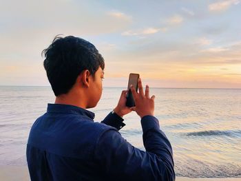 Young man photographing sea against sky during sunset