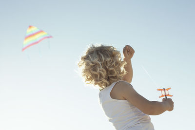 From below back view of unrecognizable cute little boy with curly blond hair launching colorful kite under cloudless blue sky on sunny summer day