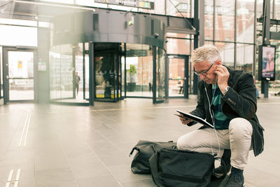 Full length of senior male commuter holding digital tablet while crouching by bags at railroad station