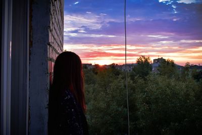 Woman standing by window against sky during sunset
