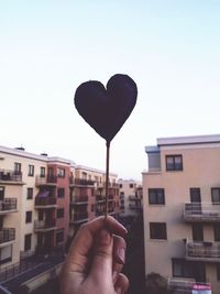 Cropped hand of woman holding black heart shape against buildings