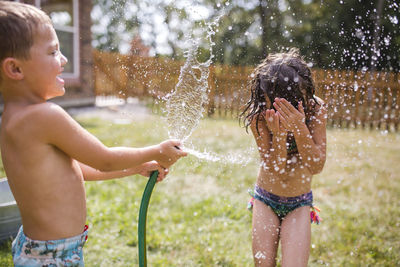 Shirtless brother spraying water on sister through garden hose at yard