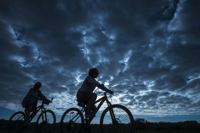 Silhouette women riding bicycles on field against cloudy sky