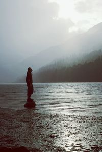 Man standing on rock at shore against mountains in foggy weather