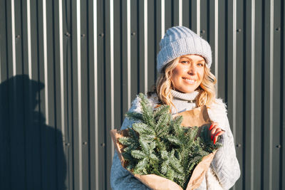 Portrait of woman in winter clothes with fir branches in hands against background of grey wall