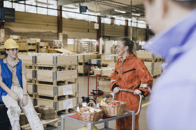 Smiling young female worker with food on push cart talking with colleague in warehouse