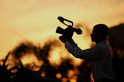 Side view of camera operator photographing against sky during sunset