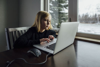 Girl using laptop computer while sitting by window at home