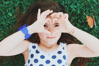 Directly above portrait of girl making heart shape while lying on grass