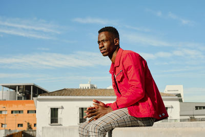 Young man in red jacket sitting on wall in city