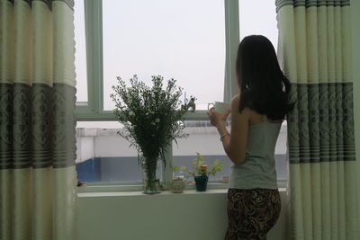 Side view of woman having drink while looking through window at home