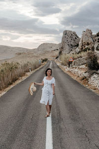 Front view of beautiful young woman in white dress standing on open road.